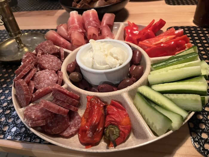 Tapas med salami, prosciutto, paprika, gurkstavar, oliver och en skål med ost, upplagt på en sektionerad tallrik.
