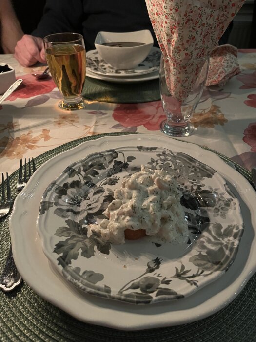 Räktoast serverad på en blommig tallrik, bredvid glas med servett och ett glas dryck på duk med blommönster.