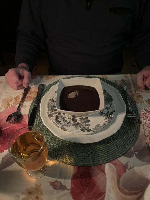 En person sitter vid ett dukat bord och har en tallrik med svartsoppa framför sig, tillsammans med ett glas dryck och bestick.