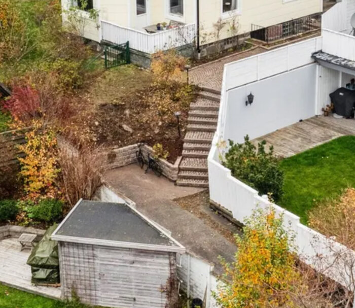 Vy över bostadstomt med trappa, uteplats, gräsmatta och träd. En cykel står lutad mot en sluttning. Ett förråd i trä finns i ena hörnet.