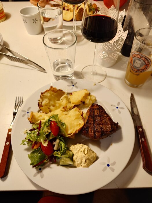 Bakad potatis med grillad rostas, sallad och bearnaisesås på en tallrik, med glas vin, vatten och öl vid en dukad middag.