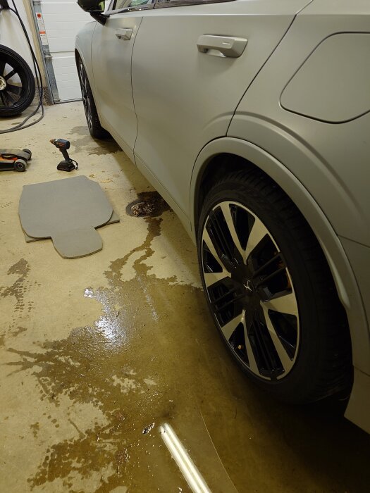 Bil med nya vinterdäck i ett garage, omgiven av verktyg på golvet, inklusive en borrmaskin och teleskopskaft. Vattenfläckar syns på golvet.