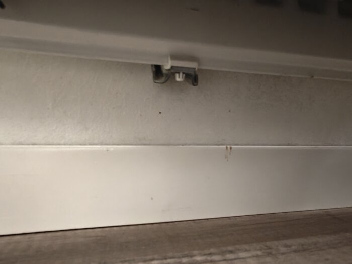 Grå väggyta med monteringsdetalj och skruv upptill, vit list nedtill samt brun golvyta.