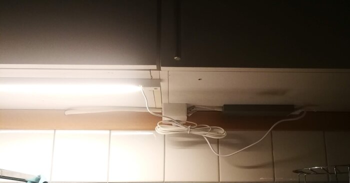Trådfri LED-ljuslist från Ikea monterad under köksskåp med inkopplad fjärrkontroll och elkabel, ovanför vitt kakel.