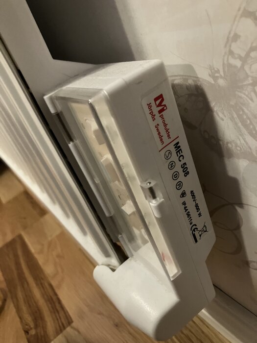 Vitt elektriskt element med trasig termostat monterad på blommig tapet och trägolv i bakgrunden.