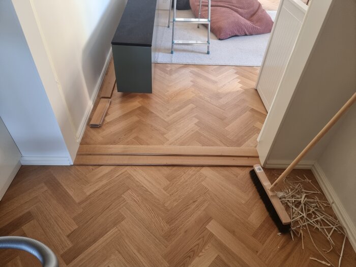 Två tröskelplattor placerade över en nivåskillnad mellan två rum med olika golv, med brädbitar och en sopborste synliga.