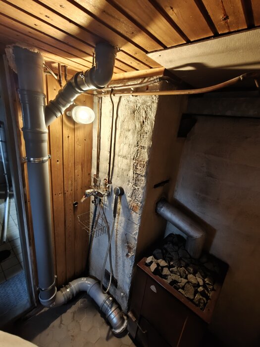 Vedeldad bastu i 50-talshus med rör isolerade med PU-skum genom taket, samt bastuaggregat med stenar och en dusch med avloppsrör i hörnet.