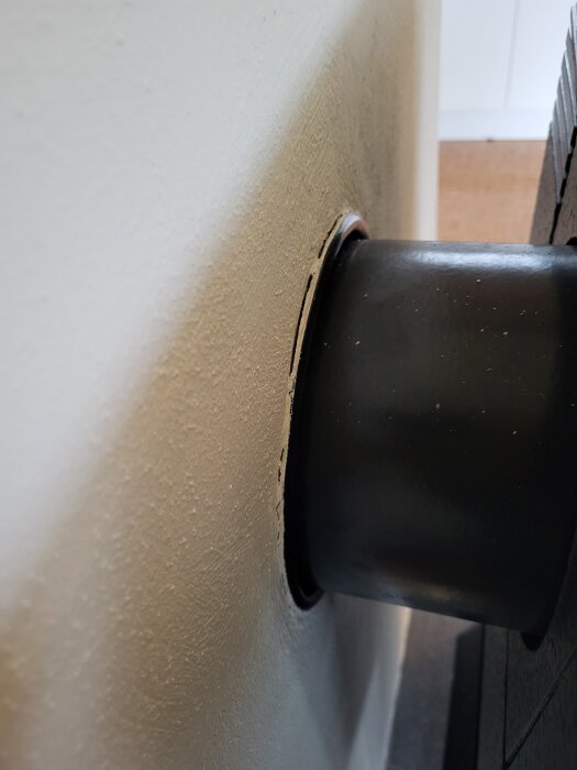 Svart röranslutning till kamin genom vitmålad vägg med synlig spricka vid kanten.