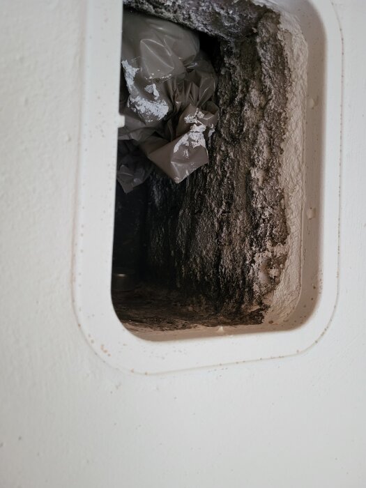 Öppning med insikt i skorstenskanal, visar mörk missfärgning och brunt papper eller plast som fyllning.