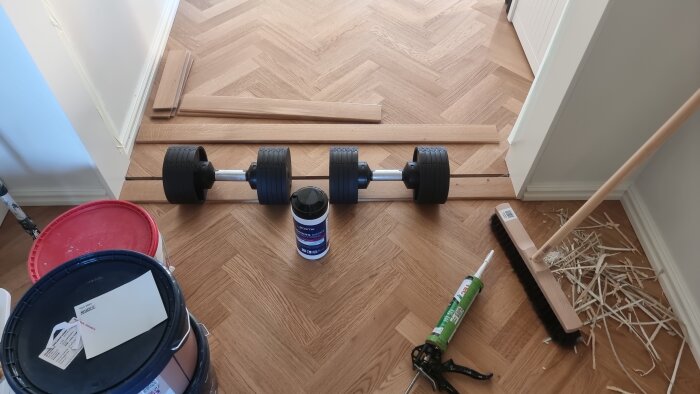 Golvlister fixeras med hantlar, omgiven av målarburkar, pensel och verktyg.