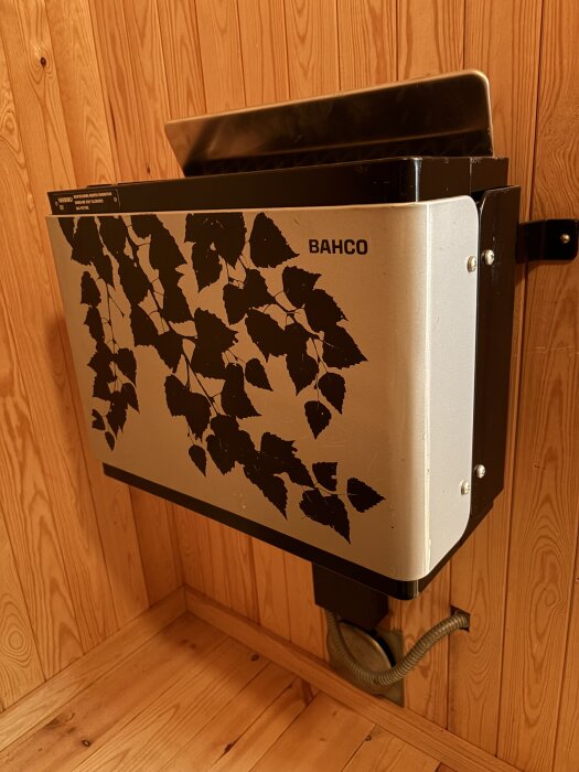 Bastuaggregat med lövdesign monterat på trävägg, märkt BAHCO, med kabelanslutning.