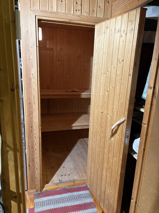 Öppen bastudörr i trä, visar ett oanvänt bastuutrymme med träpanel på väggarna och ett sittbänkssteg.