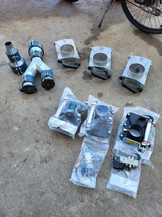 Överblivna delar från spånsuginstallation inklusive skjutspjäll och ventiler av fabrikat Dustcontrol, utlagda på marken bredvid en cykel.