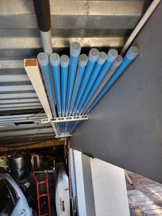 Blå alurör förvaras under ett tak, ordnade i en linje ovanför ett förråd, med en bil och annan förvaring synliga i bakgrunden.