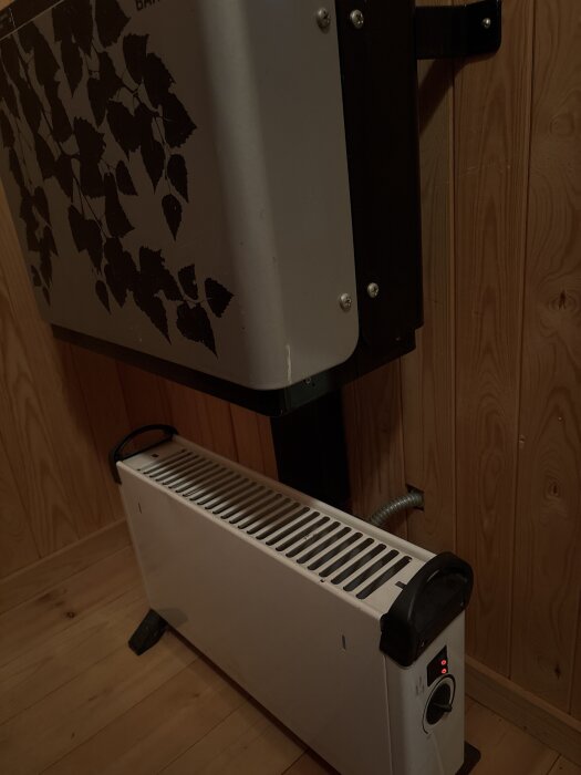 Ett vitt elektriskt element placerat mot en träpanelvägg, under en dekorerad enhet med lövmönster.