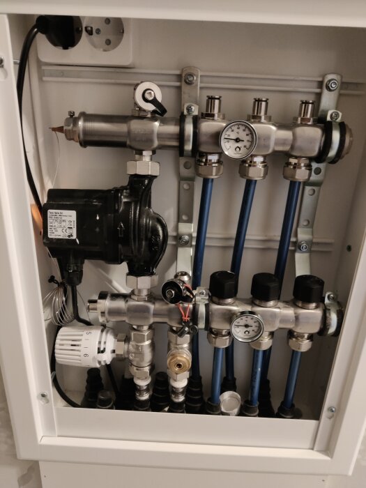 Värmefördelarsystem med rör, ventiler och två temperaturmätare uppvisar 15 grader, del av ett vattenburet golvvärmesystem i kök och hall.