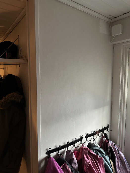 Vägg i hallen med vita gipsskivor och rad med färgglada jackor hängande på svarta krokar under ett fönster som släpper in ljus.