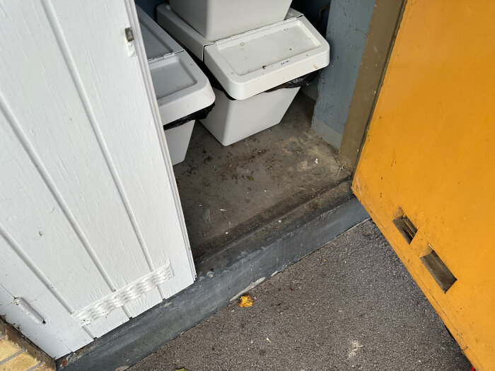 Öppet soprumsdörr med vit dörrkarm, synlig insida med två vita soptunnor på ett grått cementgolv, bredvid en gul dörr.