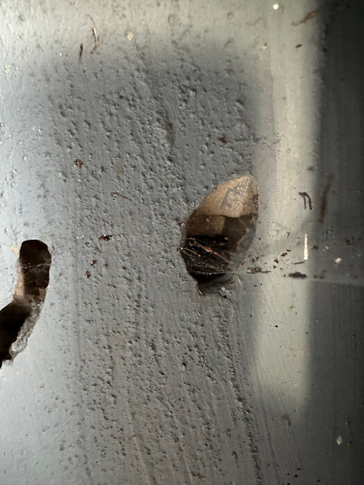 Närbild av en vägg med två hål, där ett större och ett mindre blottar insidan av materialet, vilket kan vara gipsskiva eller tunt isoleringsmaterial.