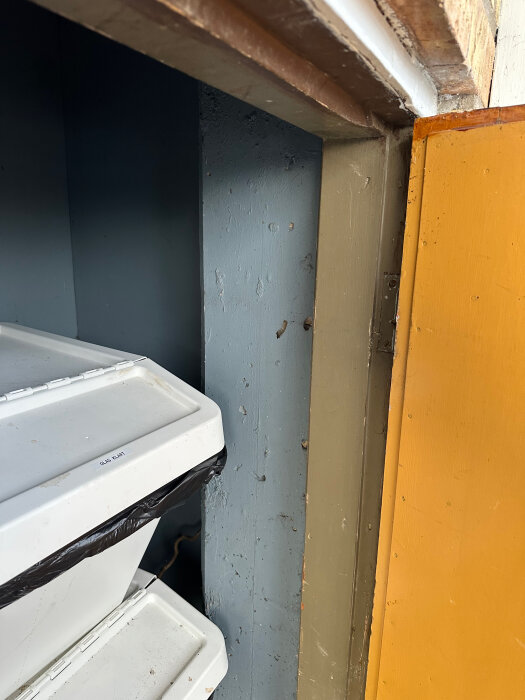 Interiör av ett soprum med två vita soptunnor, en blå och en gul vägg, samt en bred dörrkarm upptill i bild.