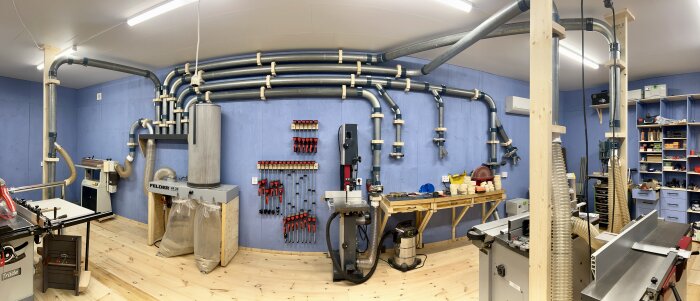 Snickeriverkstad med rör på vägg och tak för maskinanslutning, verktyg på väggen, och arbetsbänk med diverse verktyg och tillbehör.