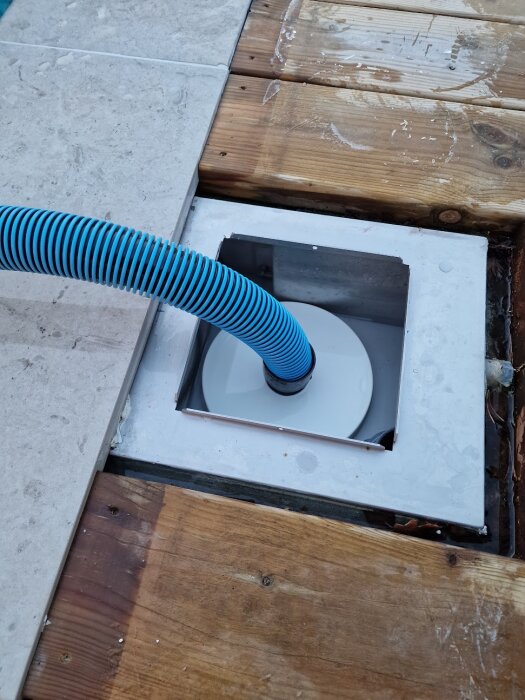 Blå slang kopplad till poolens bräddavlopp, delvis täckt av trätrall och kakel.