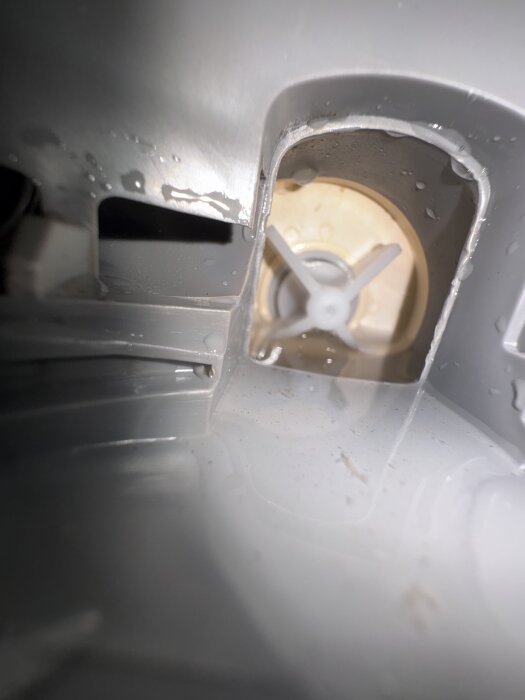 Närbild av insidan av en diskmaskin visar dräneringsområde och en gul backventil delvis borttagen, med synliga vattenrester runtom.