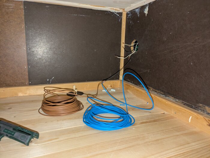 Elektriska kablar och en dosa i ett hörn av en garderob, med träytor och en spikpistol synliga på golvet.