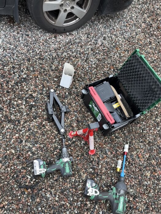 Verktyg för däckbyte inklusive en saxlyft med hexfäste, två gröna borrmaskiner och en svart verktygslåda ligger på grus bredvid ett bildäck.