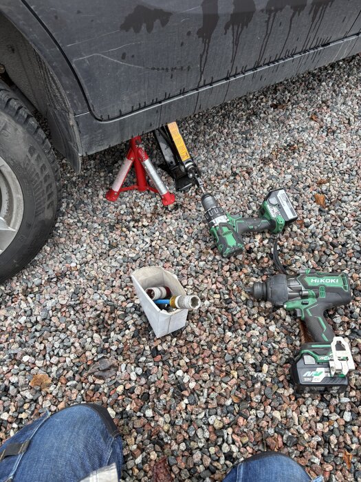 Borrmaskin och hexfäste vid bil för däckbyte, domkraft och säkring i grus. Verktygen förbättrar och snabbar upp processen.