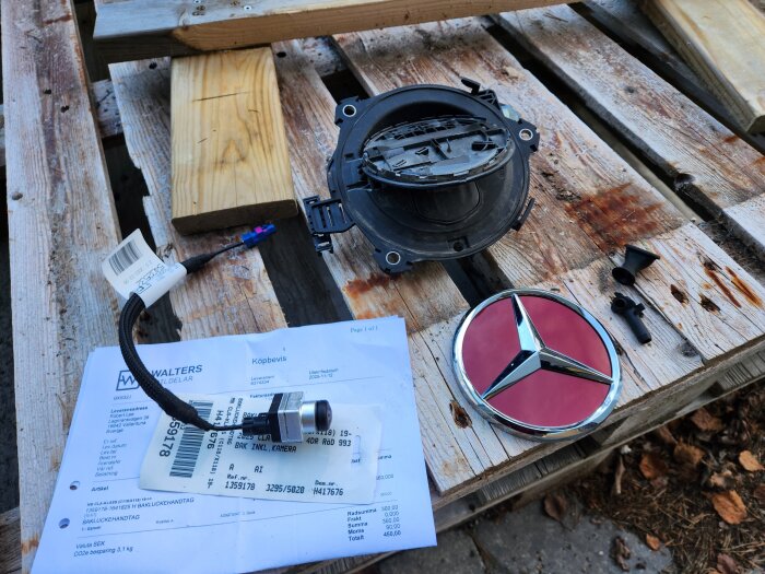 Begagnade bildelar från en Mercedes CLA-25 samt kvitto placerade på en träpall, inklusive handtagsmekanik och ett rött Mercedes-emblem.