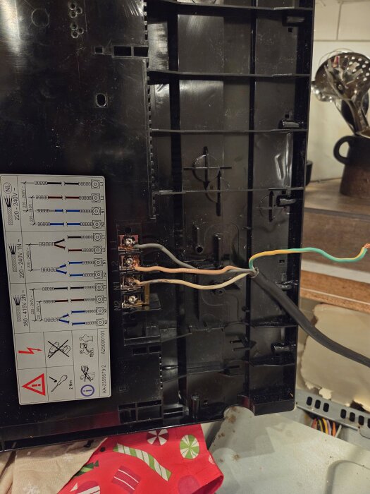Elektrisk kopplingsbox med ett kopplingsschema synligt. Tre exponerade kopparledningar: brun, blå och gul-grön. Bakgrunden visar köksredskap.