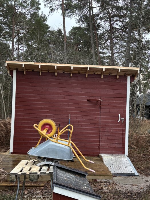 Rött skjul med förlängt takfot, omgiven av skog. Skottkärra och material ligger på bord framför.