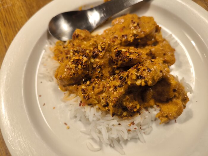 Tikka Masala med chiliflakes på ris, serverad på en vit tallrik med en sked.