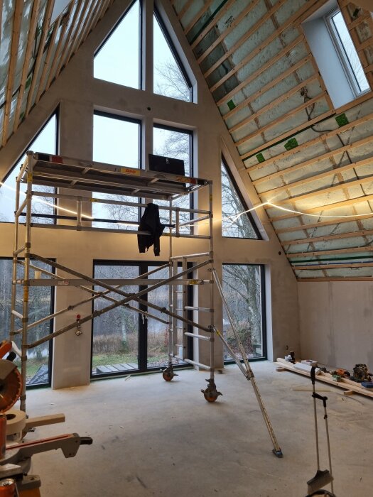 Byggarbetsplats inomhus med stora fönster och ställning för målning, osb-skivor och verktyg på golvet, rum under konstruktion.