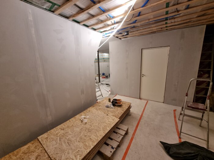 Under renovering: en hall med gipsväggar, en nyinstallerad dörr, stege och verktyg på OSB-skivor. Takhöjd synlig med rörinstallationer.