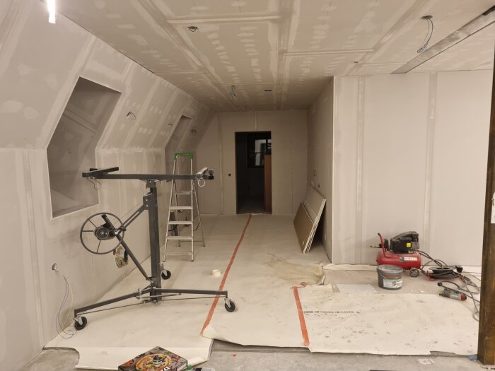 Rum under renovering med gipsväggar, tak, en stege och verktyg som skivhiss och kompressor syns.