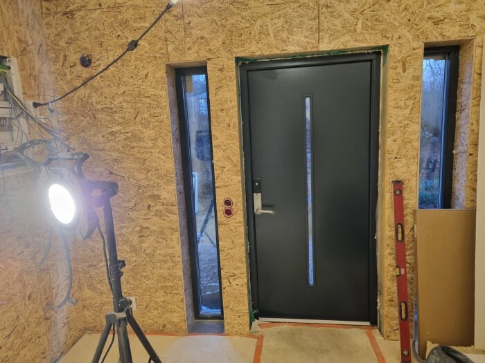 Svart dörr installerad i hall med OSB-skivor på väggarna, byggarbetslampa och vattenpass syns.
