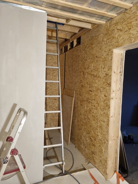 Ställning och stege i hall under renovering med osb-skivor och gips; taket är öppet och det pågår arbete.