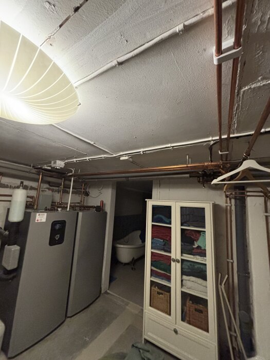 Källare med oisolerade radiatorrör i taket, bergvärmepump och skåp med handdukar, samt ett badkar i bakgrunden.