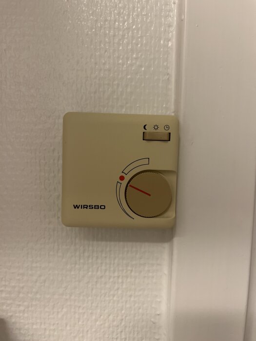 Gammal Wirsbo-termostat på väggen, beige med vridknapp och symboler för olika temperaturinställningar, del av golvvärmesystem från 90-talet.