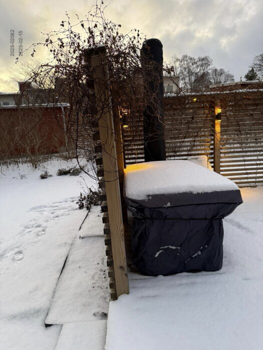 Snötäckt altan med insynsskydd och belysning, förråd och SPA-bad i bakgrunden. Spår i snön visar användning av området, vintrig bakgård.