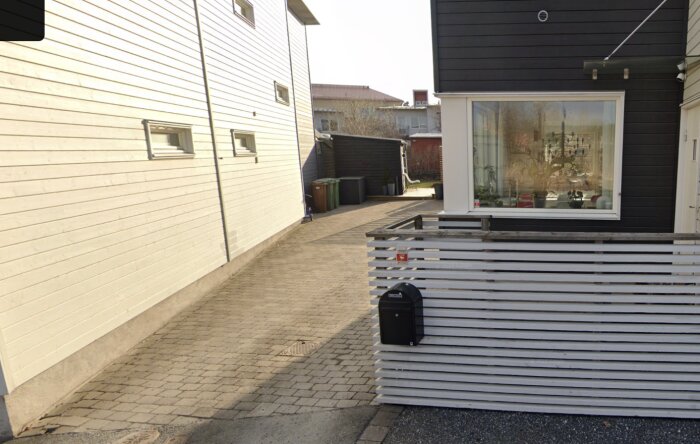 Marksten och förråd vid modern byggnad, omgivna av ljus träpanel och en vit staketdel med en svart brevlåda.