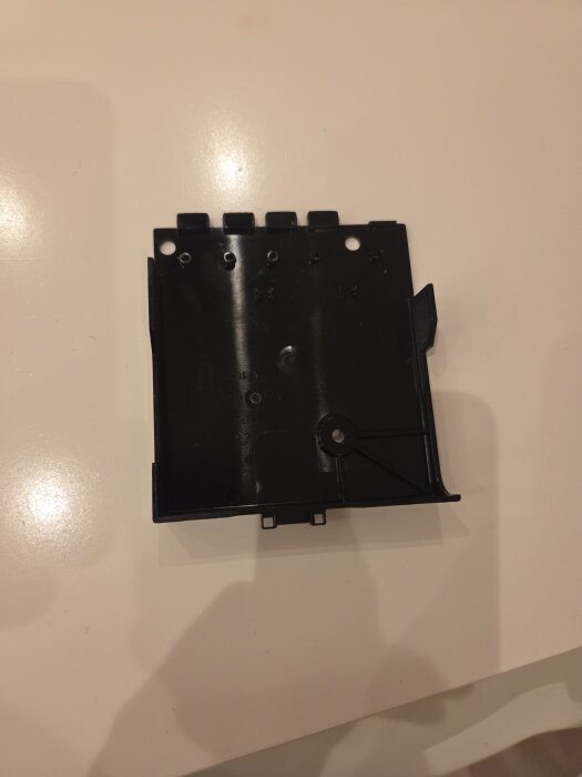 Svart plastdel med hål och klämmor liggande på ett ljusare bord.
