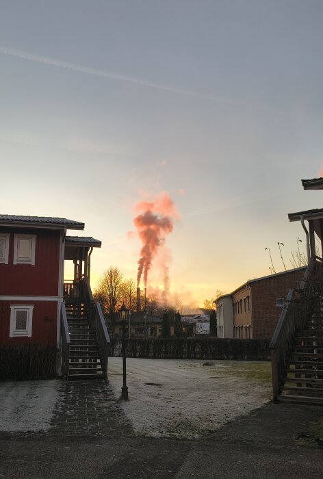 Rök stiger från fabriksskorstenar i bruksort, omgiven av byggnader vid solnedgång.