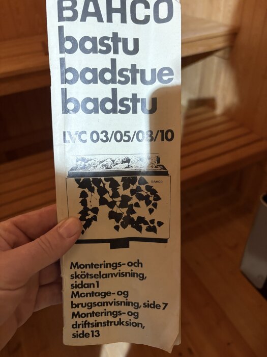 Broschyr för Bahco bastuaggregat med montering och skötselanvisning i flera språk, hålls av en hand med trähylla i bakgrunden.