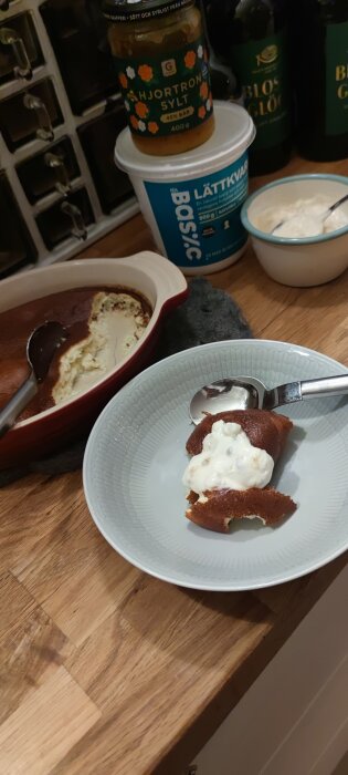 Keso-ostkaka serverad på tallrik med hjortronkvarg. Burkar med hjortronsylt och lättkvarg syns i bakgrunden på köksbänken.