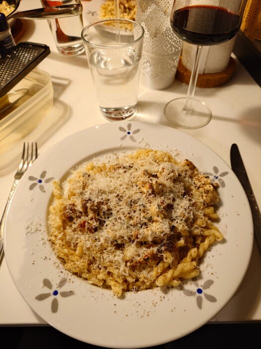 Tallrik med pasta, kyckling, soltorkade tomater och riven ost. En glas vin och vatten syns i bakgrunden.