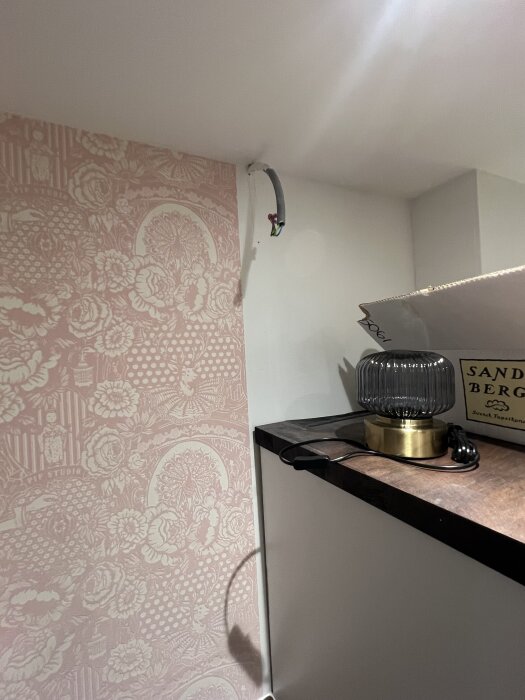 Tapetserad köksvägg med utskjutande elkabel från taket. Intill står en bordslampa på en träyta.