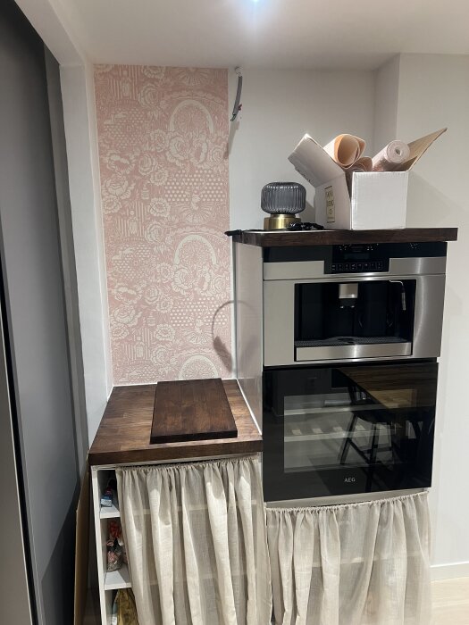 Kökshörna med rosa blommig tapet, halvmonterad elsladd vid tak, ugn, skärbräda, lampa och kartong på ovansidan samt tygdraperi.